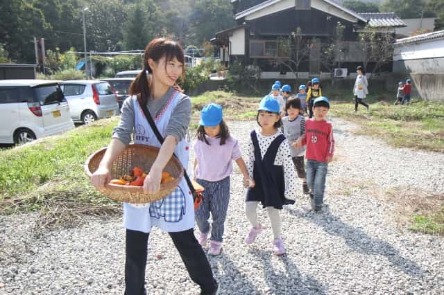 先生と手を繋いだ園児達が収穫した柿を持って列になって歩いている写真