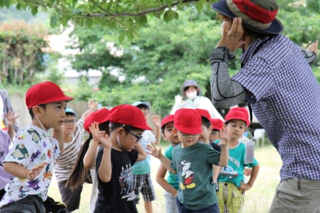 赤い帽子を被った園児達が先生と一緒に耳に手を当てて音を聞いている写真