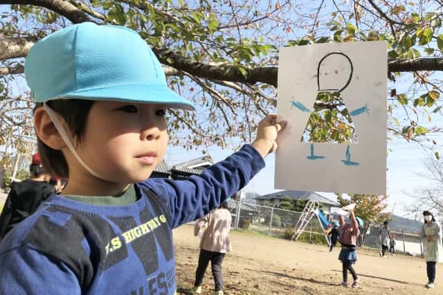 男の子が人が描かれた絵（服の部分には穴が空いている）を木の葉に当てがって木の葉を服の代わりにしている写真